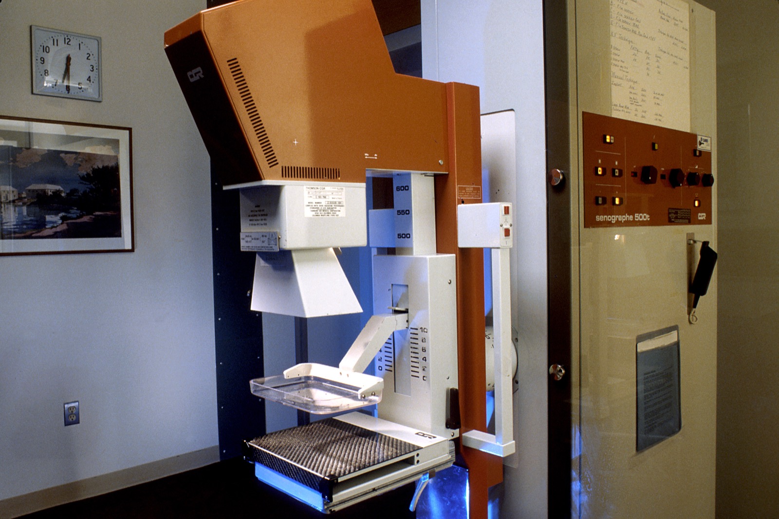 A mammography unit in a clinic exam room, the compression paddle and detector below the X-ray head.